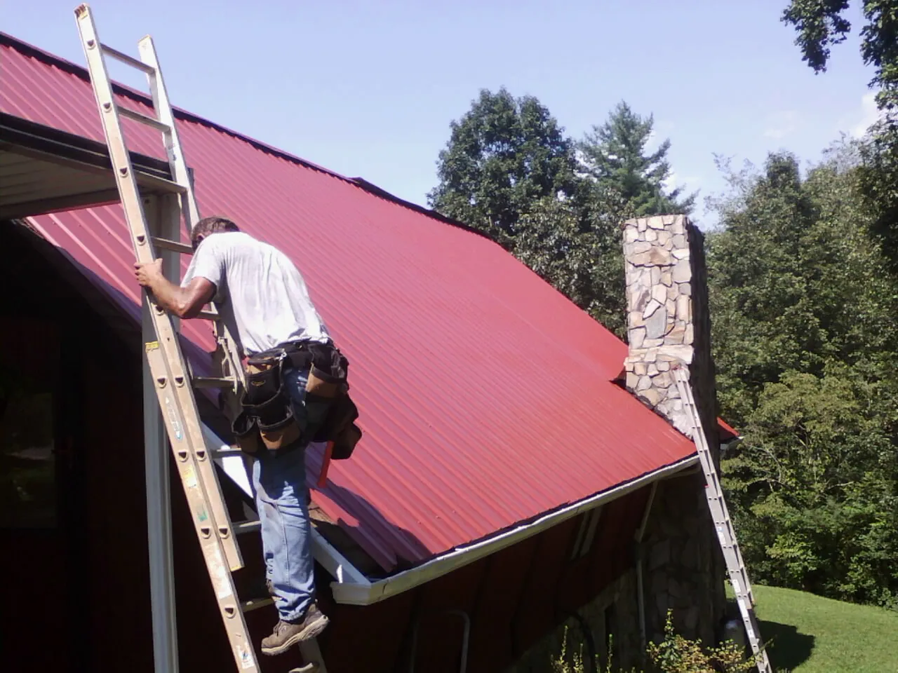 Roofer on a ladder beside a red metal roof and stone chimney — Soffit & Fascia Repair by Roofing Brothers in Blain